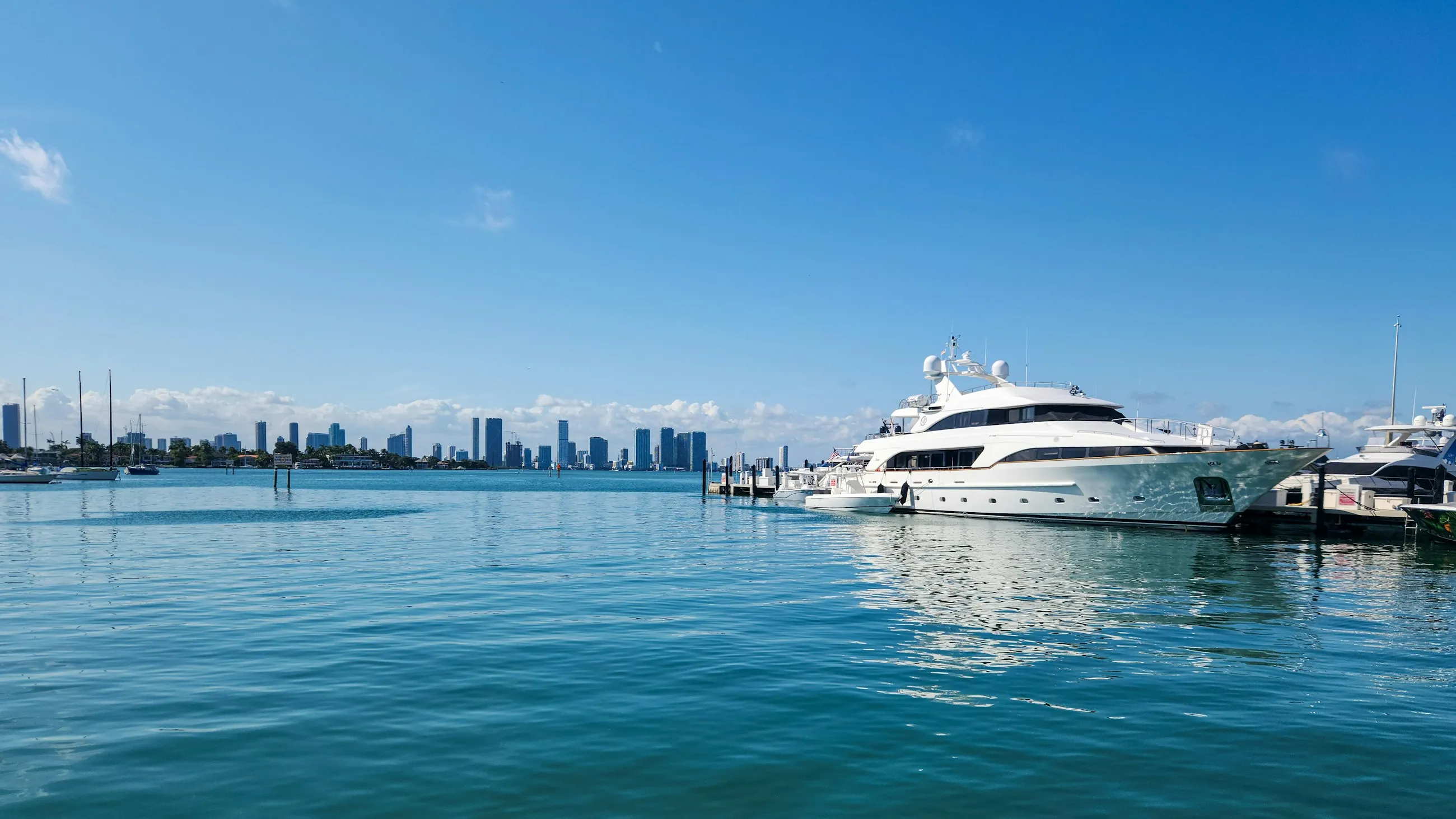 A white luxury yacht moored on calm turquoise water, with the Miami skyline in the distance